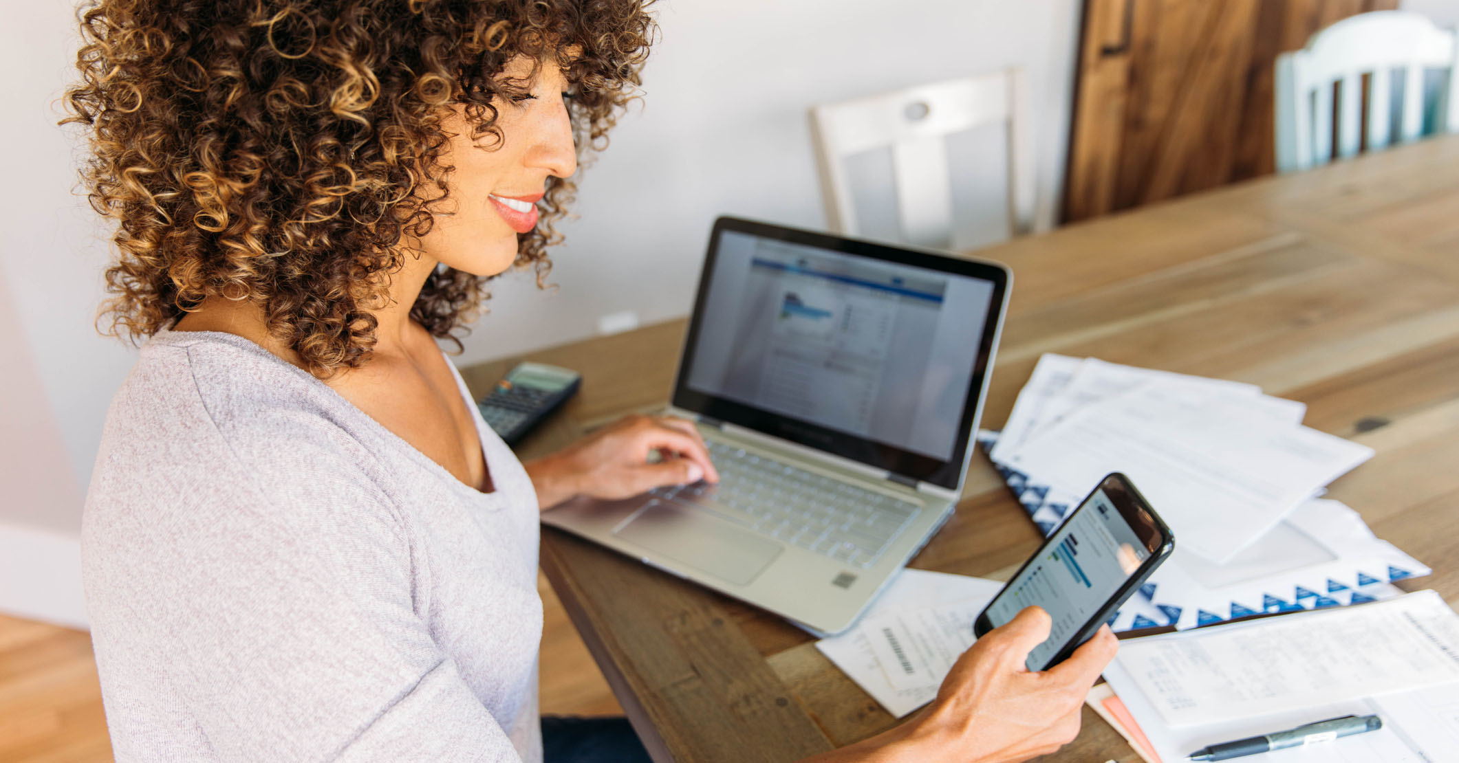 A woman sits at her dining room table with laptop and financial reports doing her monthly budget. She is smiling at the ease of use as she works on her smart phone banking app to do monthly finances, pay taxes and save money for the future.